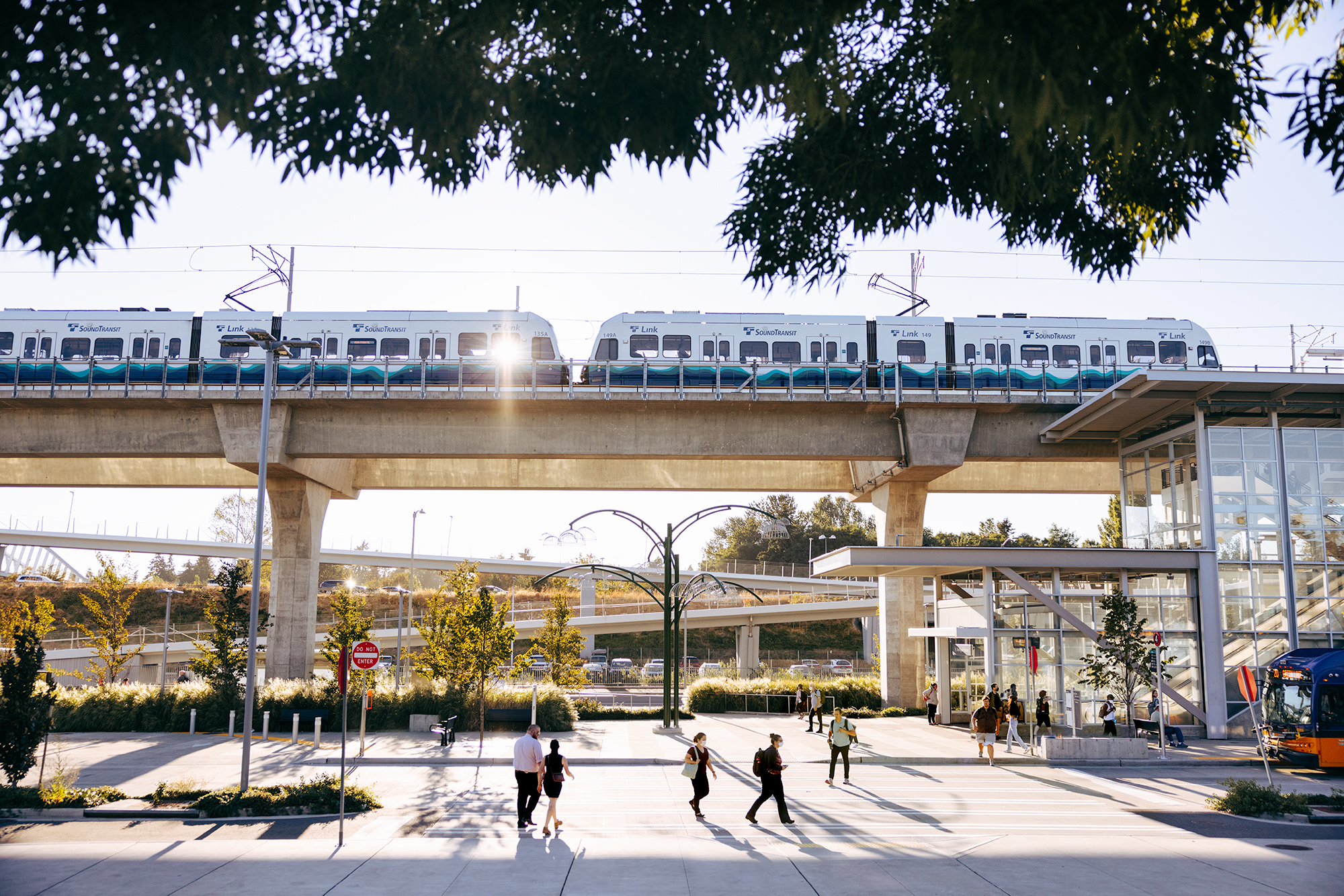 A Link light rail train approaches an elevated station as pedestrians and bus connections move about on the ground-level.